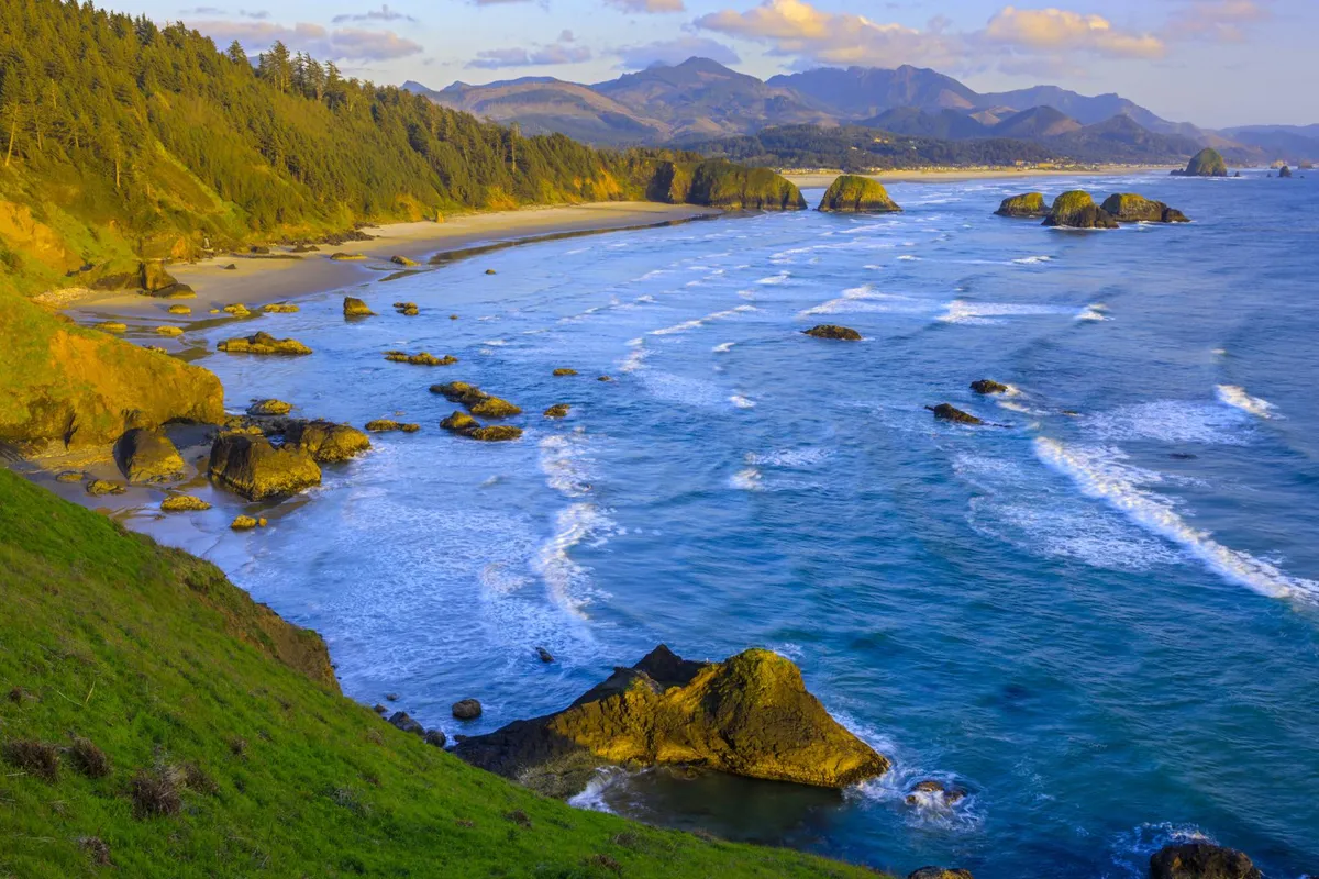 A breathtaking view of a rocky shoreline stretching into the distance, with gentle waves lapping at the rocks and distant mountains visible on the horizon. The setting is tranquil, inviting, and embodies the natural beauty of the Oregon Coast, perfect for exploring with pets.