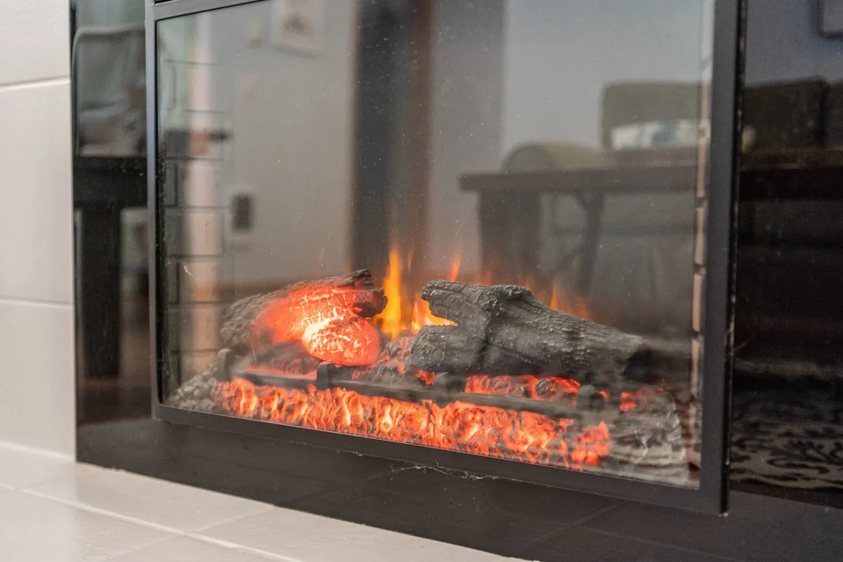 A close-up of a glowing electric fireplace with realistic flames and warm lighting. The scene suggests a comfortable indoor space perfect for relaxing, reading, or sharing stories together on cool evenings.