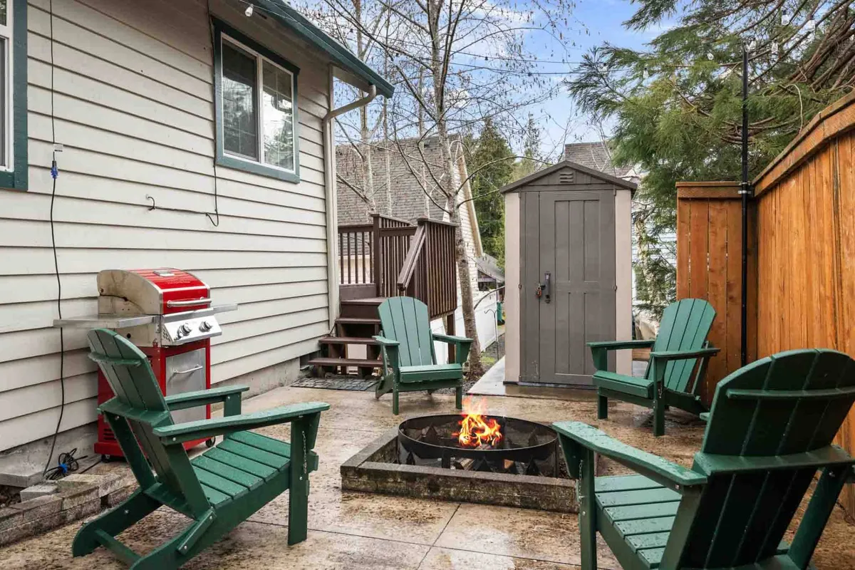An exterior view of a fenced backyard, complete with a fire pit, Adirondack chairs, grill, and visible storage shed. The enclosed setting offers both security and relaxation, making it perfect for pets and outdoor leisure.
