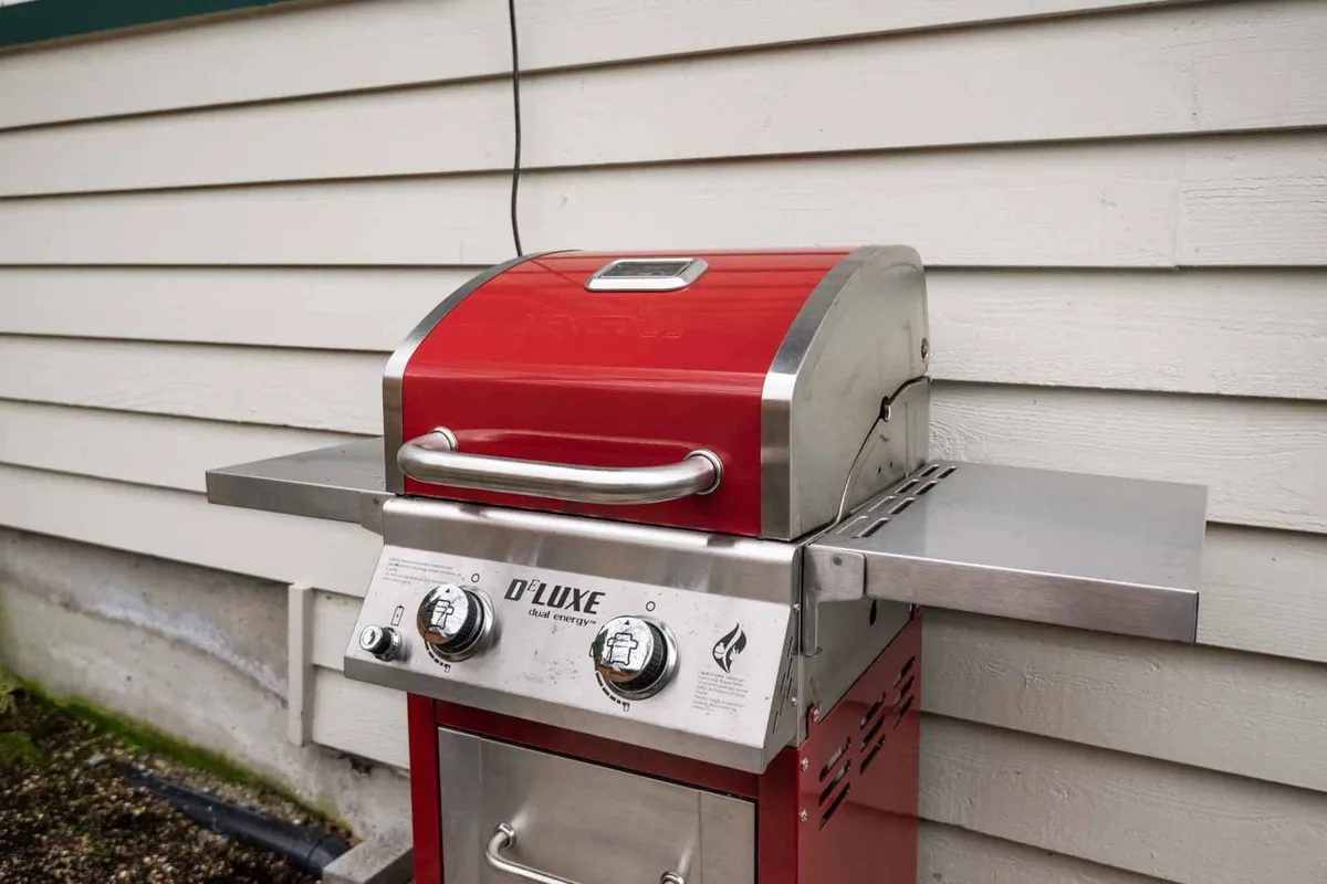 A welcoming outdoor patio scene featuring a vibrant red gas grill set against a backdrop of greenery. The patio area offers ample space for gatherings, with enough room for guests to mingle and enjoy the fresh air, surrounded by trees that create a secluded, forested atmosphere.