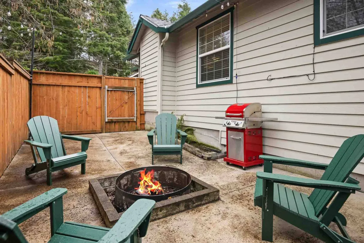 This image presents an inviting exterior setup featuring a fire pit surrounded by Adirondack chairs, with a grill nearby. The area is shaded by mature trees, creating a cozy, natural retreat ideal for relaxing, gathering, and enjoying the outdoors.