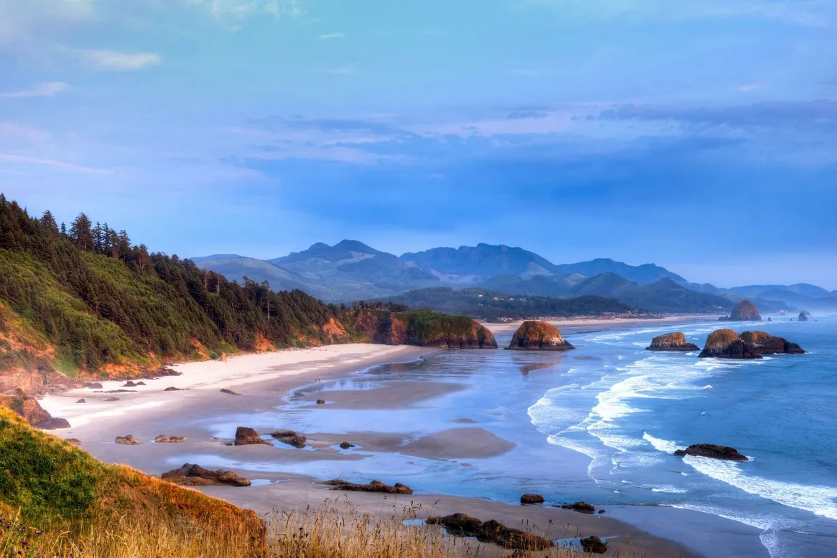 A wide, serene coastal view with rugged coastline, sandy beach, distant mountains, and waves gently crashing ashore. The setting is perfect for beachcombing, sunset watching, and exploring tide pools, capturing the region’s unspoiled beauty.