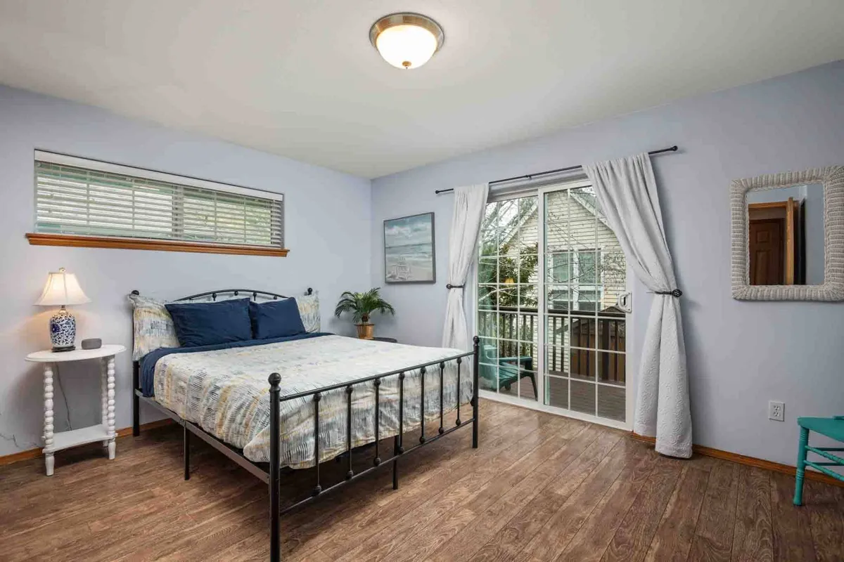 A bedroom with patterned bedding, a sliding glass door that opens to a balcony, a decorative lamp on the bedside table, and a potted plant. The room is filled with natural light, offering a serene and airy feel.