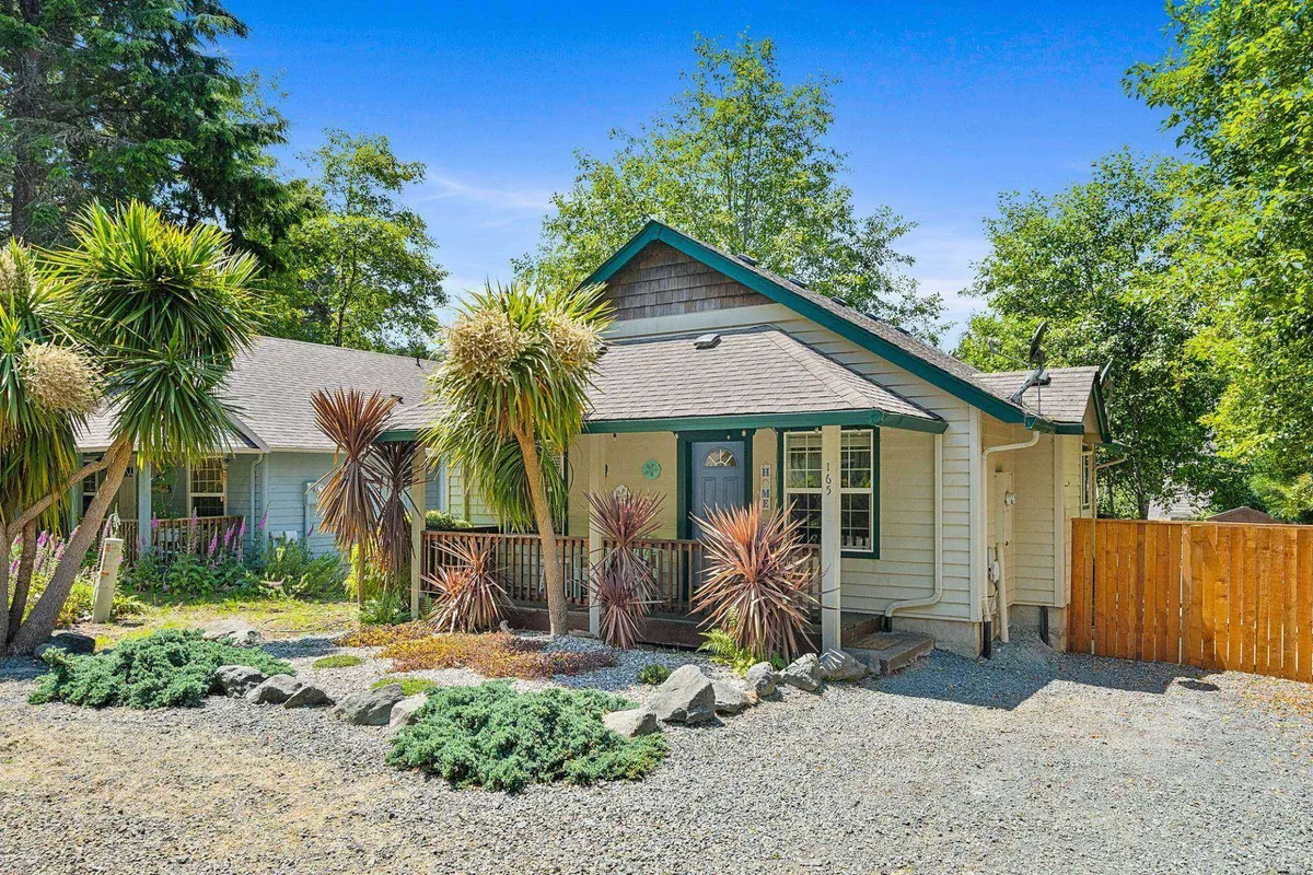 The image shows the welcoming front porch of a charming single-level bungalow, surrounded by lush tropical plants and a quaint stone pathway leading to the entrance. A wooden fence frames the property, creating a peaceful and private atmosphere perfect for relaxing. The golden hues of sunset bathe the porch, hinting at the tranquil evenings and inviting mornings guests can enjoy outdoors.